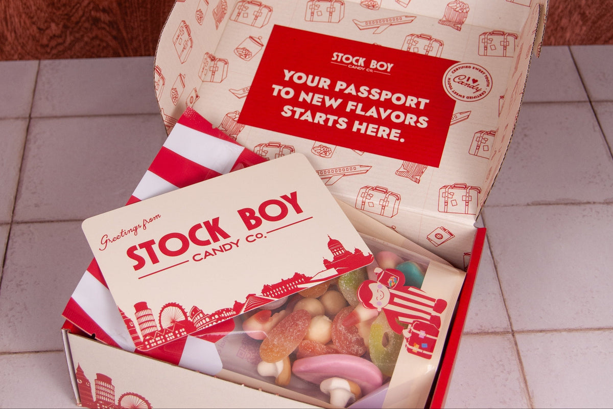 An open Stock Boy candy gift box on a light tiled surface against a dark wood background. The box contains a branded paper bag and a clear window showing assorted colorful candies.
